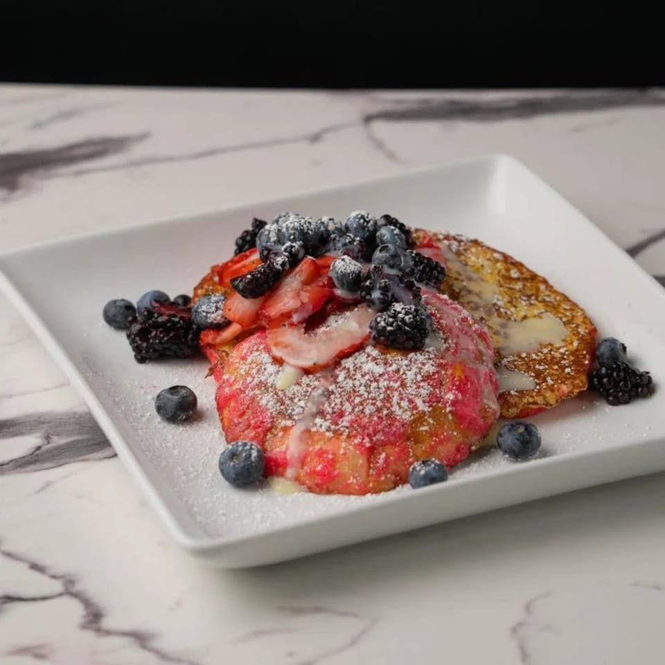 Two berry-topped pancakes, one red velvet, on a white plate with powdered sugar and syrup.