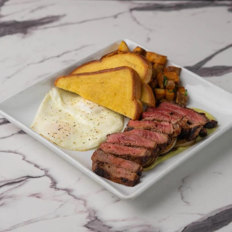 Steak and eggs breakfast with toast and roasted potatoes on a white rectangular plate.
