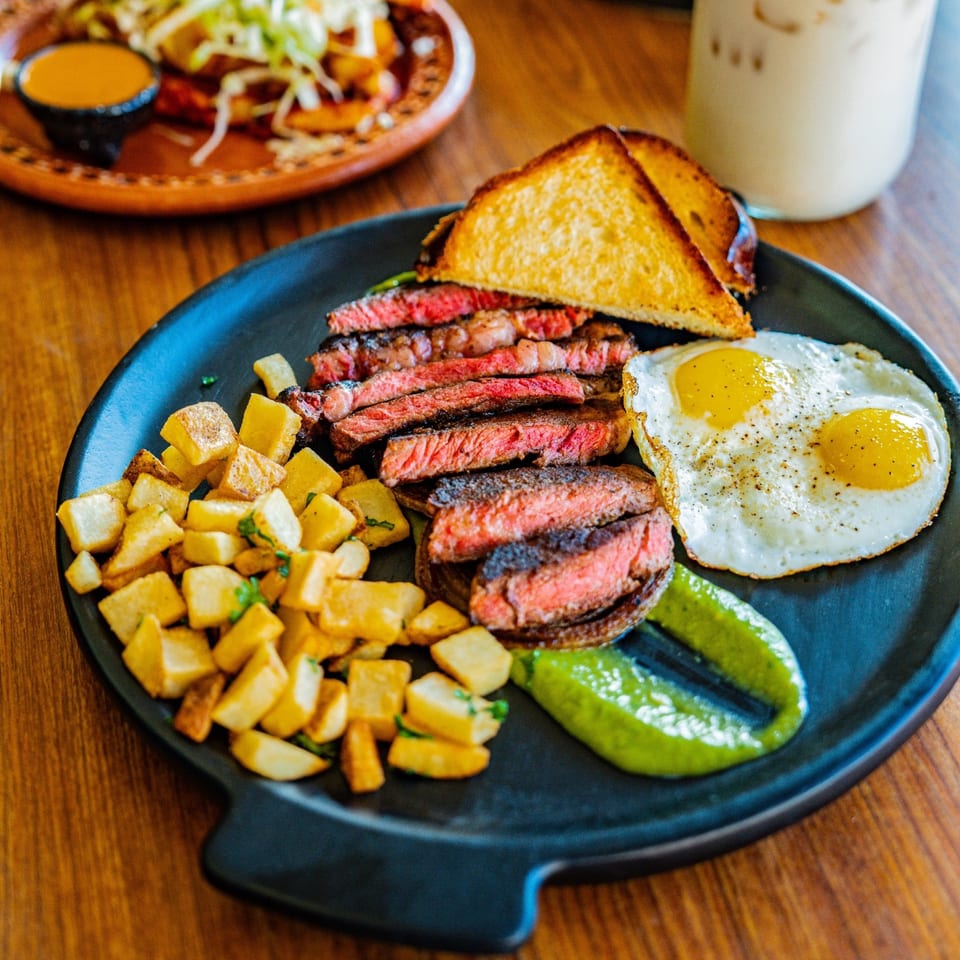 Steak and eggs breakfast with home fries, toast, and green sauce on a black plate.