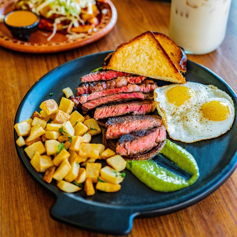 Steak and eggs breakfast with home fries, toast, and green sauce on a black plate.