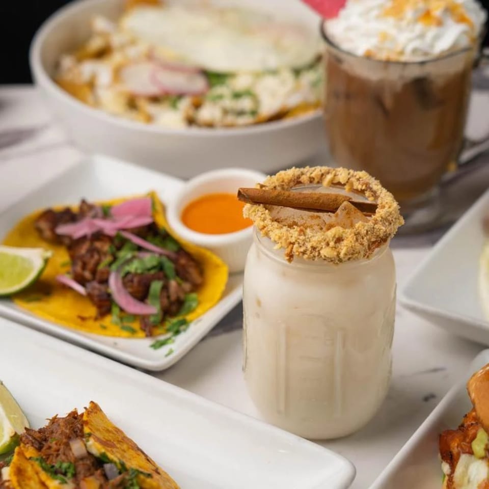 A horchata with a cinnamon stick and a taco with lime, alongside other dishes and drinks.