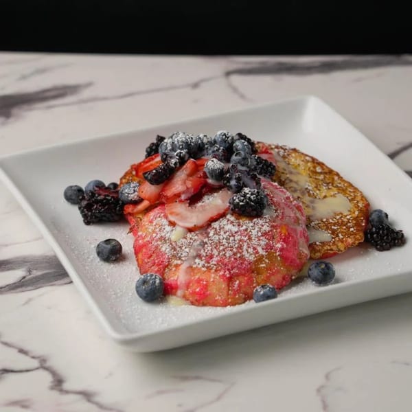 Two berry-topped pancakes, one red velvet, on a white plate with powdered sugar and syrup.